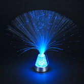 Use only descriptions that match the image shown: Fibre Optic Sensory Lamp with illuminated strands; LED fibre optic lamp with crystal-filled base; Fibre Optic Sensory Lamp glowing blue; Close-up of sparkling optical fibre tips; Illuminated crystal-filled lamp base; Remote control for the Fibre Optic Sensory Lamp; Fibre Optic Sensory Lamp displayed in a living room; Colourful fibre optic mood light in a sensory space.