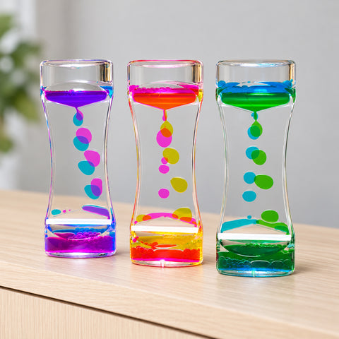 Use only descriptions that match the image shown: Lava Flow Sensory Desktop Toy with colourful bubbles; Clear acrylic liquid-motion sensory toy; Complementary coloured droplets flowing through the Lava Flow toy; Lava Flow Sensory Desktop Toy on an office desk; Liquid-motion toy in a study space; Lava Flow Sensory Desktop Toy retail displayer of 12; Individually boxed Lava Flow Sensory Desktop Toy.